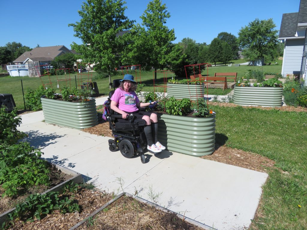 Angie alongside the fully in use Vego Garden raised beds. Showing she has full access to them at the height of her power wheelchair. 